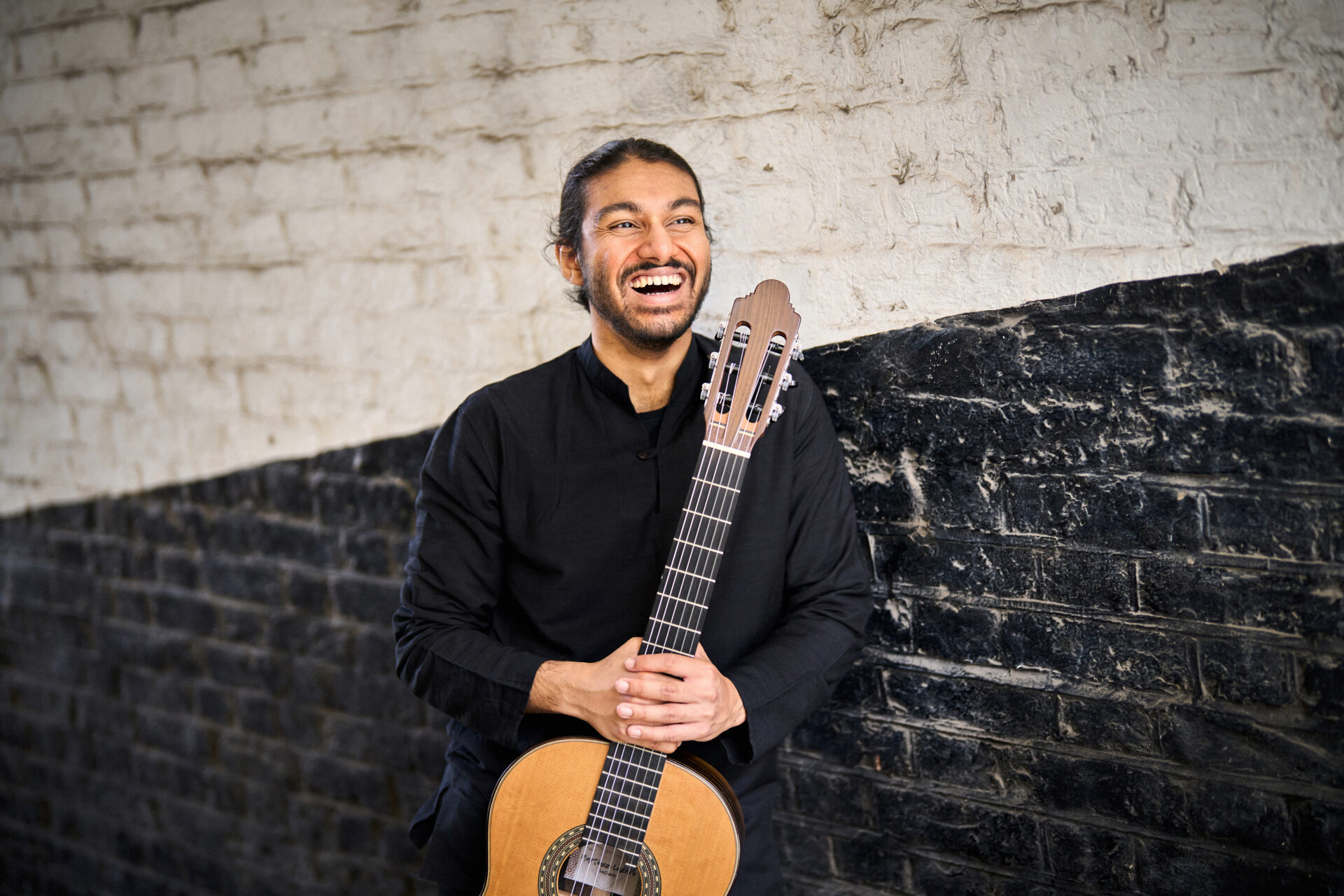 Samrat Majumder holding his classic guitar by its neck. He is laughing whilst looking off to his left. The background is a black and white brick wall, with an approximately 25 degree line separating the two colours. 